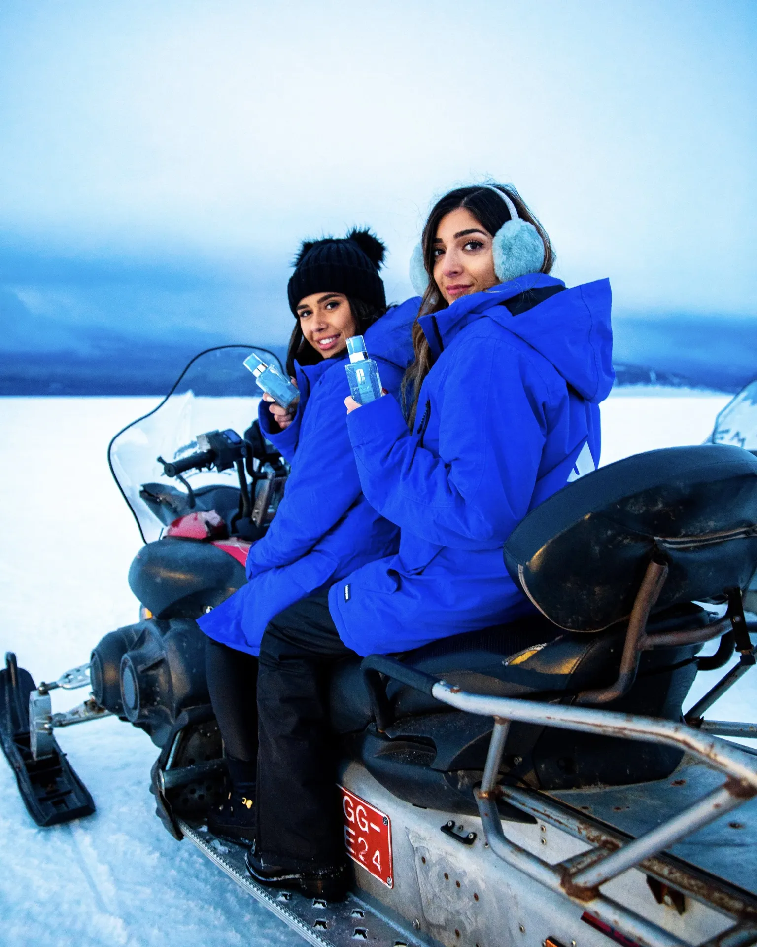 Two women in blue winter jackets and ear muffs sitting on a snowmobile in a snowy landscape, holding bottles and smiling