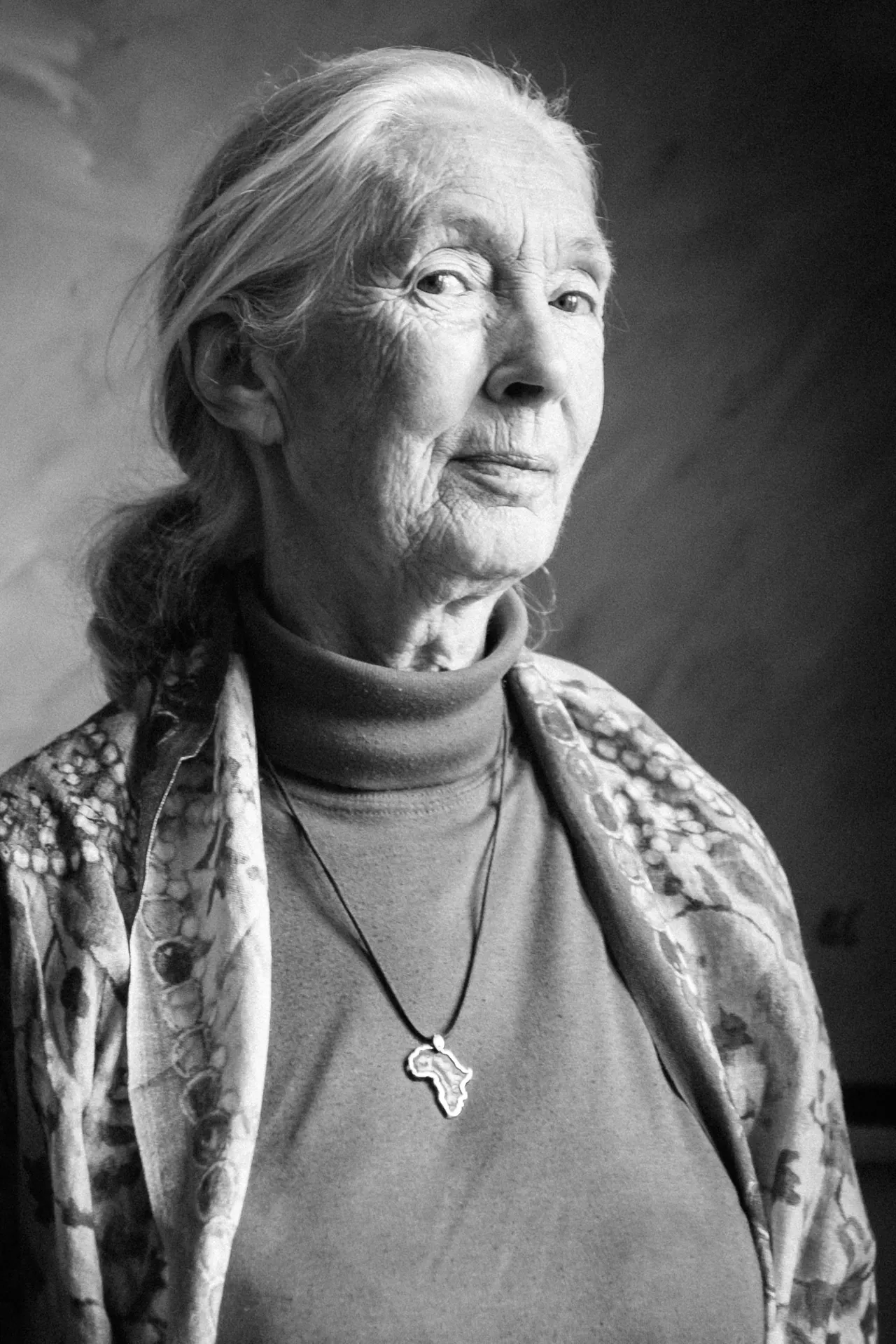 A black and white portrait of Dr Jane Goodall wearing a floral jacket, a turtleneck and a necklace with an African continent pendant