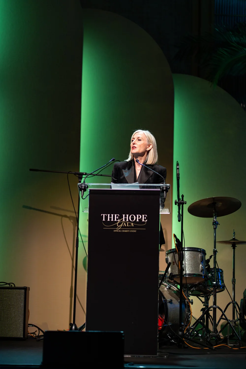 A speaker at the lectern on the stage of The Hope Gala, beside a sparkle drum kit under green stage light
