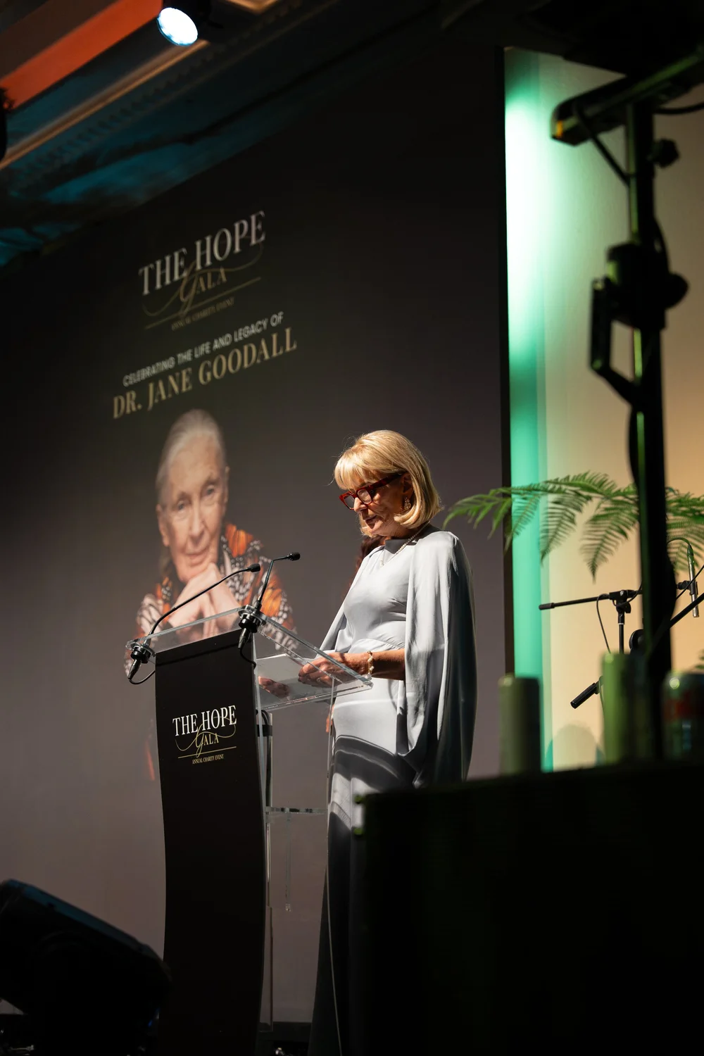 A speaker reading from notes at the lectern on the stage of The Hope Gala, in front of the backdrop portrait of Dr Jane Goodall