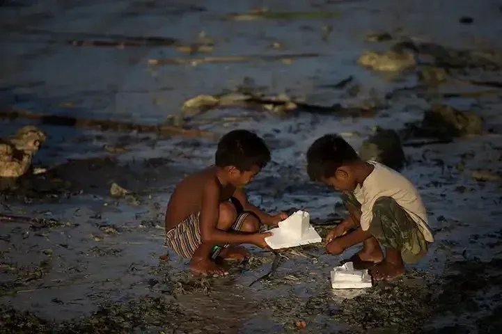 Two young boys playing with a small white object on a muddy, rocky shoreline