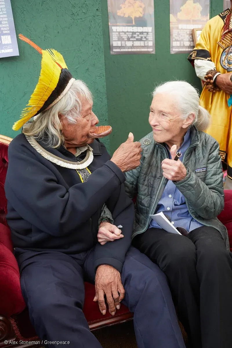 An elderly Native American man in a traditional feathered headdress beside Dr Jane Goodall, smiling and giving a thumbs up