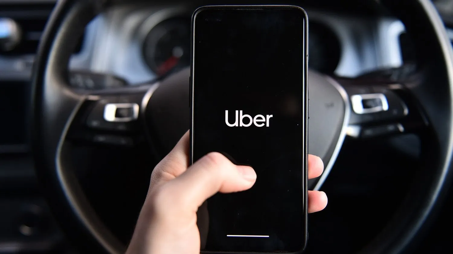 A hand holding a phone showing the Uber app at a steering wheel